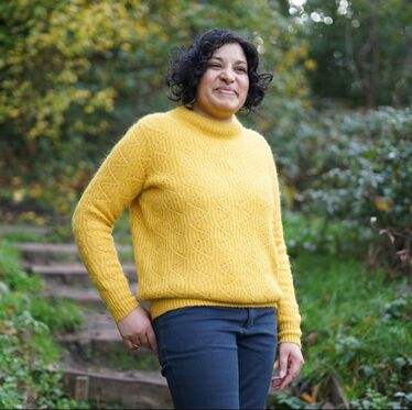 Image Description: Anuradha Kowtha, wearing a yellow sweater smiles warmly, stairs leading into a line of trees in the background.