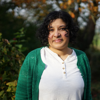 Image Description: Anuradha Kowtha, an Indian diasporic person, wears a green cardigan and smiles warmly, foliage in the backround.